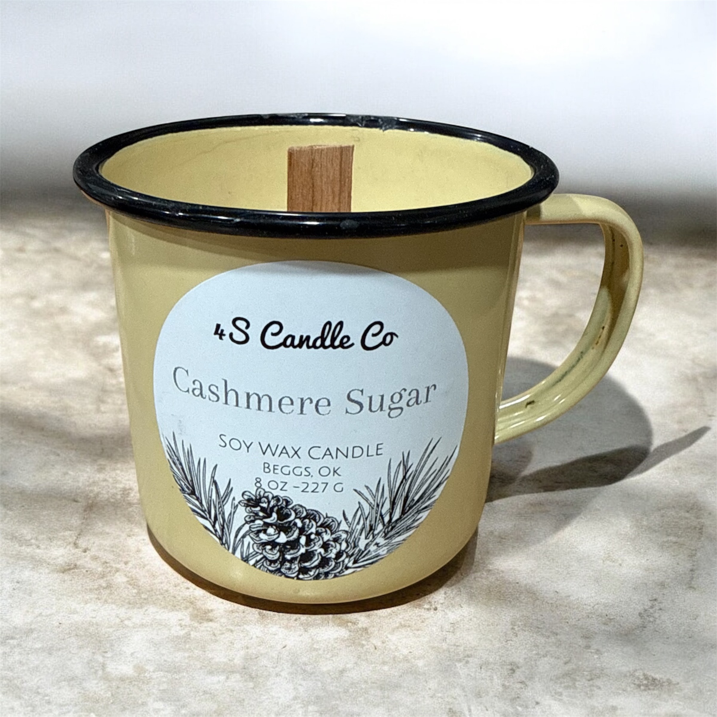 An antique butterscotch yellow camp mug is filled with white wax and a wood wick is centered. The 4S Candle Co winter label with pinecones and leaves are on the bottom of label and candle information is printed above this grapghic with candle weight, product name, location made, scent name and company name.