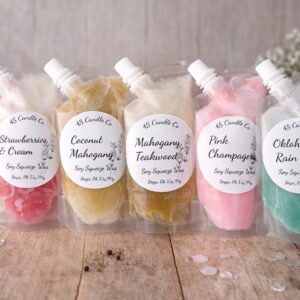 A line of 5 wax melts sit ontop of a wood table. Each melt is a differnt color and each has a label nameing the 4S Candle Co informtion including scent names. A bowl of strawberries, flowers, and a towl sits in the background.