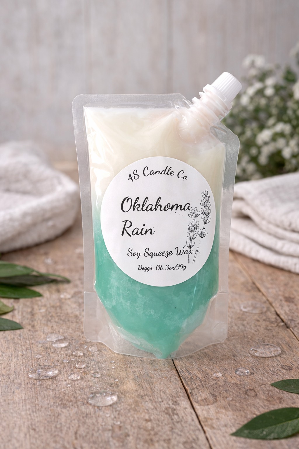 Plastic pouch with a spout that is capped to the right of the bag. Bag is filled with aqua and white wax. The melt bag sits on a wood table with water drops and green leaeves placed around the table top. White babys breath flowers is in the background.