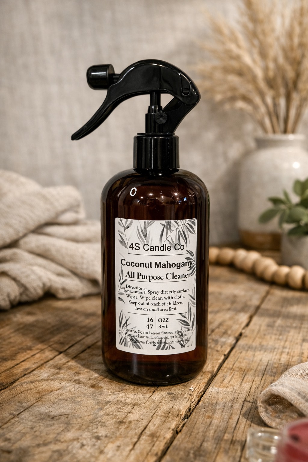 One plastic spray bottle is sitting on a wood table with a rustic indoor background design. The spray bottle has a spray nozzle. The bottle has a brown string wrapped around the top with a bow. The lable has product information consisting of company name, product name (All Purpose Cleaner) , scent name, directions, wight, place made, and warning label.