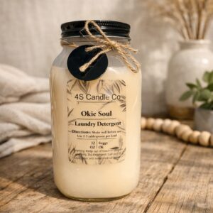 A clear plastic mason jar with a black screw on cap sits on a rustic wood table. The jar has a brown string wrapped around the topped with a bow and a black circle charm attached. The jar is labed with a clear label wich gives product information of weight, product name, instrictions, warning, company name, and scent name. The background consists of a white wall, a blanekt, beads, and flowes in a vase withc areall blurred in the backdrop of the setting.