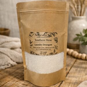 brown craft bag with a clear window showing shite powder laundry detergetn. The package has a clear label with product information: 4S Candle Co, Southern Mesa, INstructions of how to use and weight. (32oz). Package sits on a rustic wood table and background has a plant in a vase, beads, and a blanket for decor.