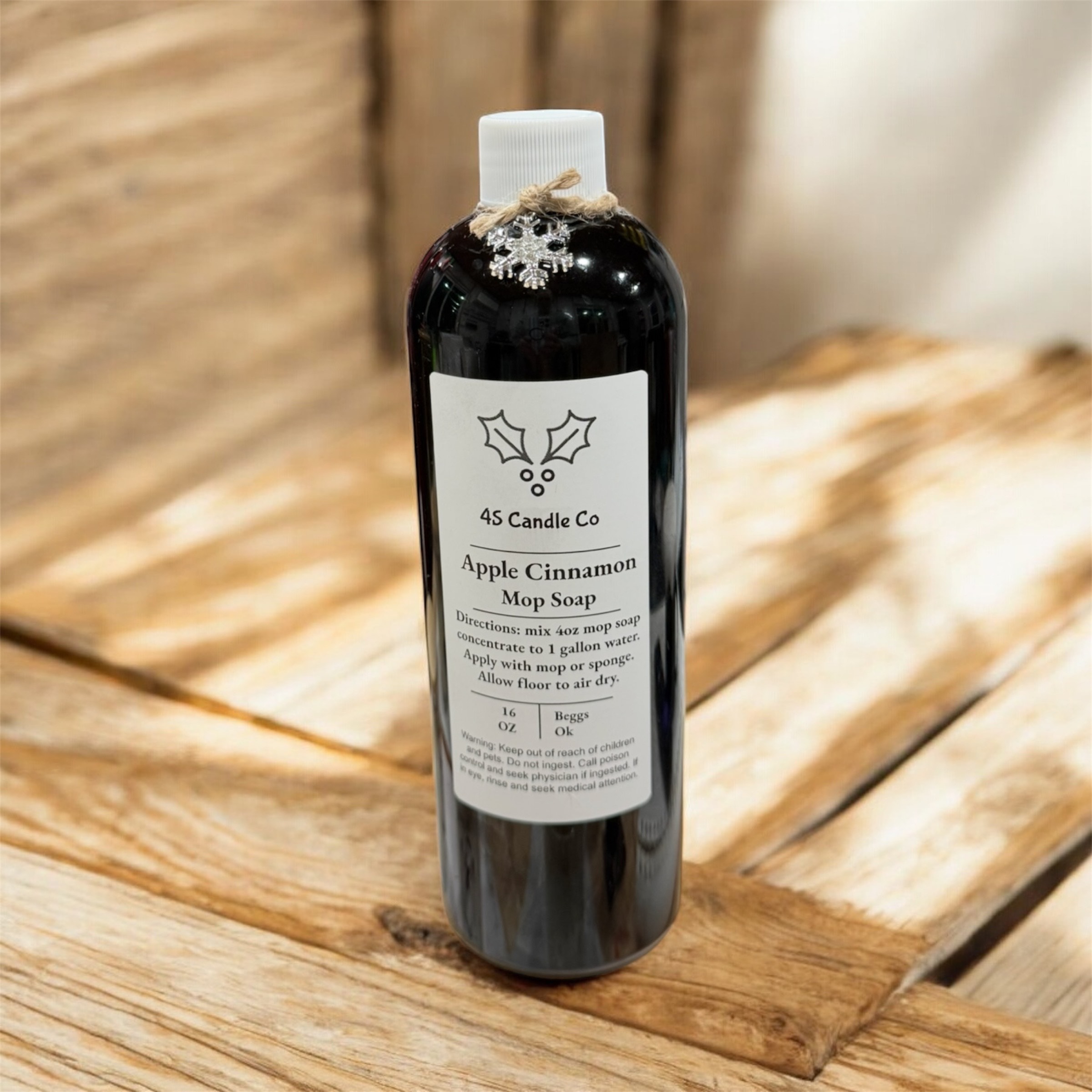 Black Bottle wtih a 4S Candle Co label placed in the center sits on top of a wood crate. The bottle has a white cap and a string with a snowflake charms wraps around the top. The background is of rustic wood and neutral colors.