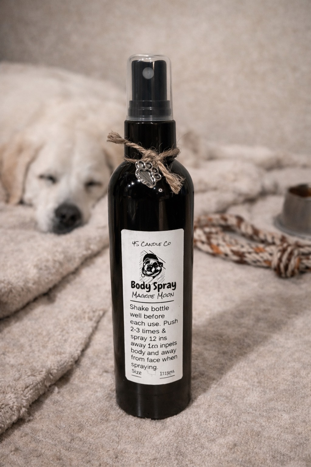 a black misting spray bottle with a clear cap, a string tied around the top of the bottle with a silver paw print attached. The bottle is 4oz in sixe, it has a label with 4S Candle co printed on it, a picture of an english bulldog, the product name: Body Spray, the name Maggie Moon and directions of how to use the product. The bottle sits on a tan rug with a partical picture of a rope, a dog bowl, and a dog sleeping in the background.