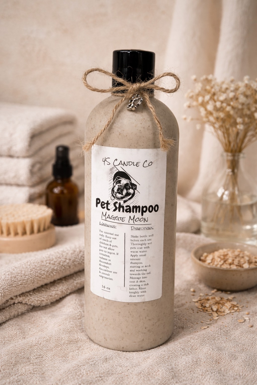 A 16oz tan bottle stis ontop of a tan towl. The bottle has a black cap and a string tied in a bow is attached around the top of the bottle with a silver paw print charm. A white label centers the bottle with the 4S Candel CO name printed on the top, a picture of an english bulldog, the product name of Pet Shampoo and Maggie Moon, Directions and a warneing lable are also on the lable. THe background consists of stacked tan twols, flowers in a vase, a bowl and a brush.