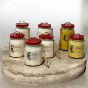 Seven differnt sized clear jars with red metal lid and handle sits ontop of a rustic concrete tabel top. Each jar has a string wrapped around the top of the jar that holds a wooded spoon. The jars are filled with white or yellow powder laundry detergents. Each jar has a label with 4S Candle Co product information. A neutral white background adds to the photo.