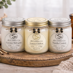 Three 6oz mason jars grouped together on a wooded circular tray. Two candle jars have a silver scre on lid and one has a gold screw on lid. Each candle jar is tied with a brown string around the top and a black bow charm attached. each candle has a 4S Candle Co lable with weight, wax type, and scent. Background cosists of rustic decor of vase, towl, mathces, plant, and blanket.