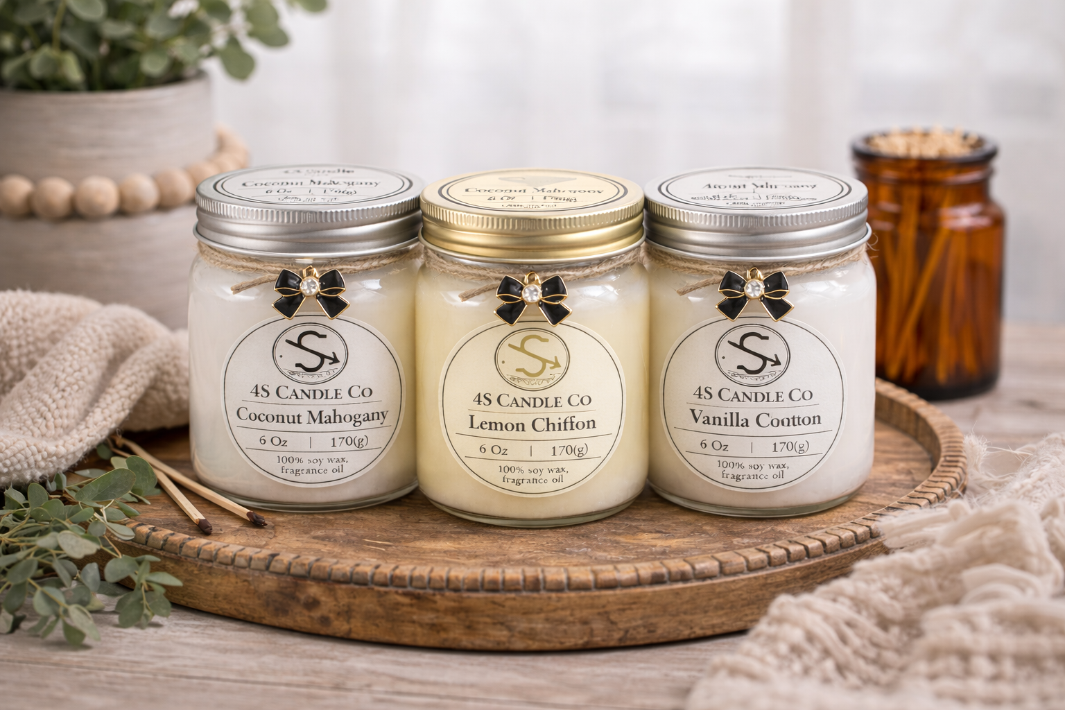 Three 6oz mason jars grouped together on a wooded circular tray. Two candle jars have a silver scre on lid and one has a gold screw on lid. Each candle jar is tied with a brown string around the top and a black bow charm attached. each candle has a 4S Candle Co lable with weight, wax type, and scent. Background cosists of rustic decor of vase, towl, mathces, plant, and blanket.