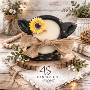 mini cow head shaped black dough bowl filled with white wax and two wood wicks placed in the center. A sunflower is attached to the top left of the cow head and a brown rustic bow is arapped around the candle. Backgrouond is rustic and has flowes, leaves, and plant decor