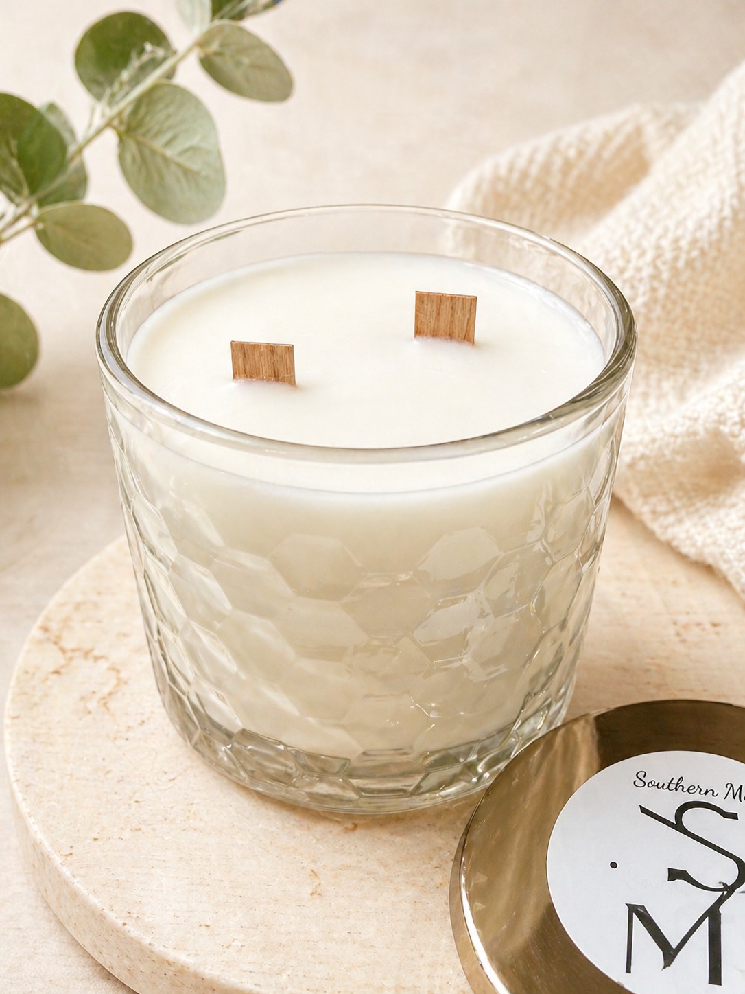 Clear candle with a hexagon pattern pressed into the glass leaving a textured effect. the candle is stitting on a granet surface ontop of a table with greenery and a blanket in the background. The candle vessel is filled with white wax and two wood wicks are centered. Particle picture of a shiny gold lid with a $ S Candle Co info sticker sits on the granet to the side of the candle.